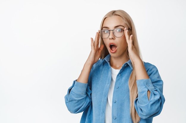 Imagen de una joven rubia que se puso anteojos y miró sorprendida a la cámara sorprendida al ver algo parado sobre fondo blanco