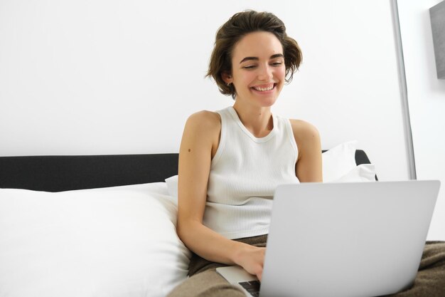 Foto imagen de una joven sonriente y feliz en el dormitorio sentada en la cama con una computadora portátil riendo y mirando