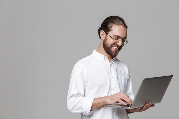 Imagen de un joven sonriente optimista guapo barbudo posando aislado sobre la pared gris con gafas usando la computadora portátil.