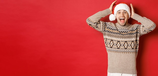 Imagen de un joven sorprendido y feliz con sombrero de Papá Noel y suéter de Navidad, recibiendo un regalo increíble, parado emocionado contra un fondo rojo.