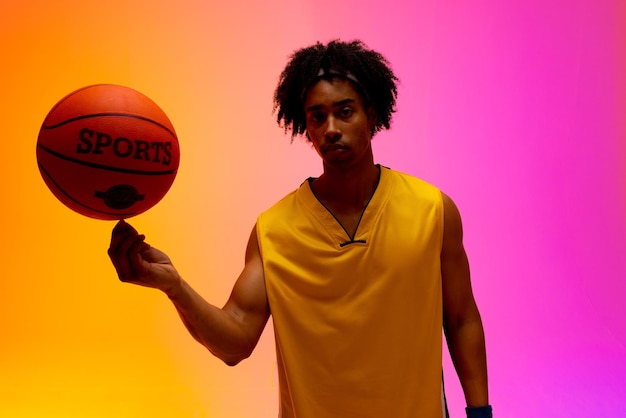 Imagen de un jugador de baloncesto birracial girando baloncesto sobre un fondo de color rosa a naranja. Concepto de deportes y competición.