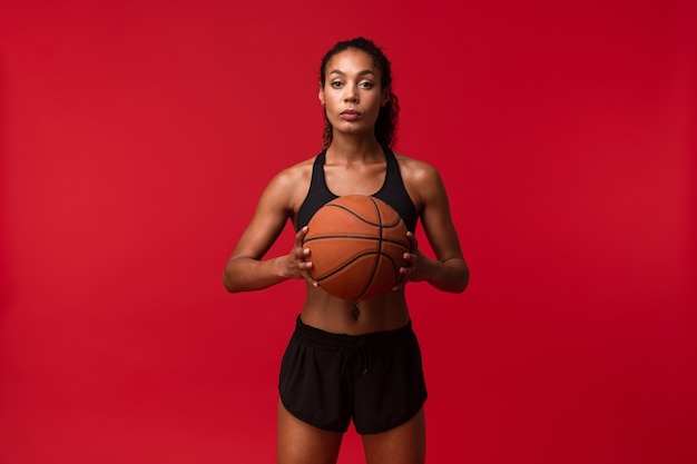 Imagen de un jugador de baloncesto de la mujer de la aptitud de los deportes africanos joven fuerte que presenta aislado sobre la pared roja que sostiene la bola.