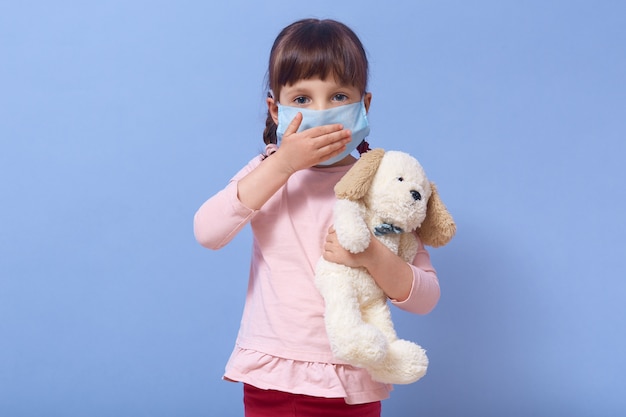 Imagen de linda chica europea en edad preescolar viste máscara médica y sostiene un juguete para perros en la mano, cubre la boca con la palma