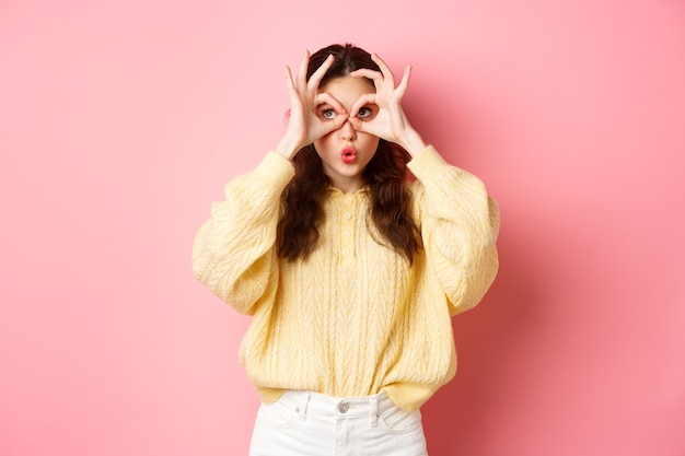 Foto imagen de una linda chica sorprendida, mirando a través de binoculares de mano, mirando a la esquina superior izquierda y diciendo wow, de pie sobre una pared rosa.