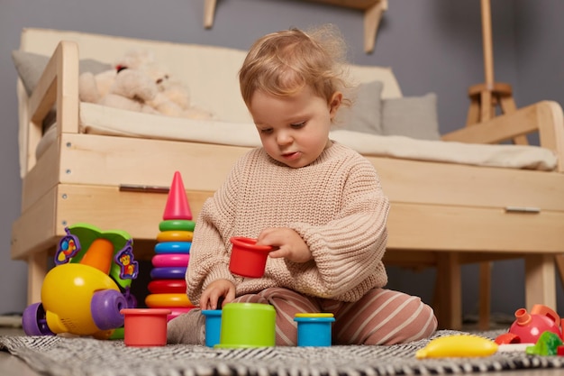 Imagen de una linda y encantadora niñita jugando con juguetes coloridos en casa sentada en el suelo jugando juegos de educación temprana aprendiendo colores y tamaños con expresión concentrada