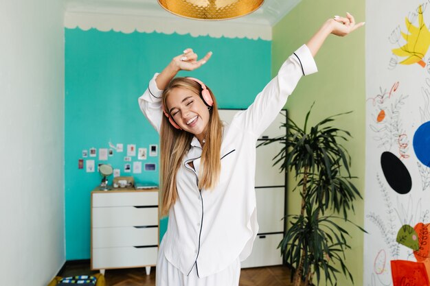 Foto imagen de una linda joven feliz en pijama en casa escuchando música con auriculares bailando.