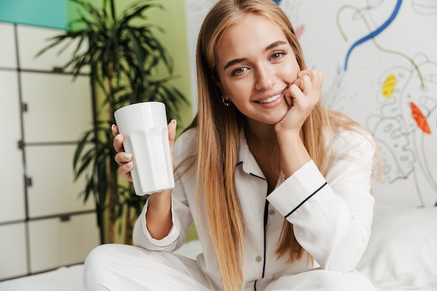 Imagen de una linda joven sonriente en pijama en casa en la cama tomando café.