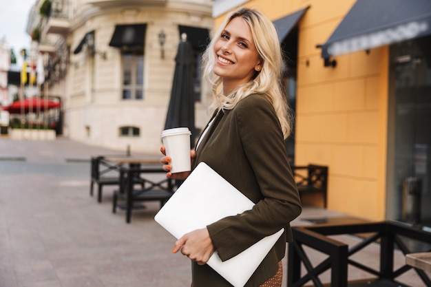 Imagen de linda mujer de 20 años con chaqueta sosteniendo café para llevar y portátil, mientras camina por las calles de la ciudad