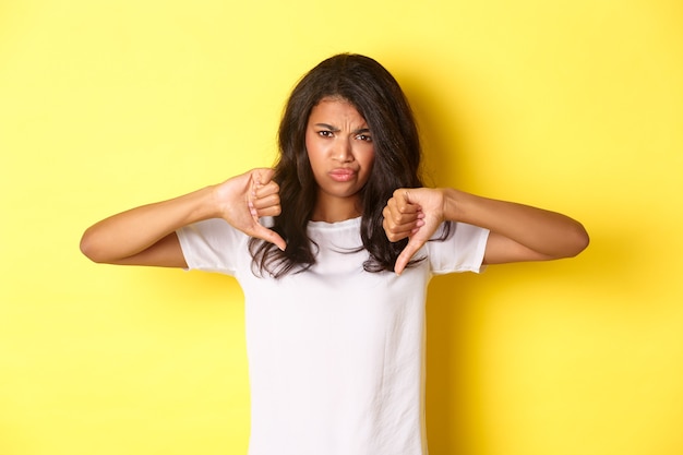 Imagen de una linda niña afroamericana escéptica y sin gracia, mostrando el pulgar hacia abajo y haciendo muecas, juzgando algo malo, de pie decepcionada sobre un fondo amarillo.