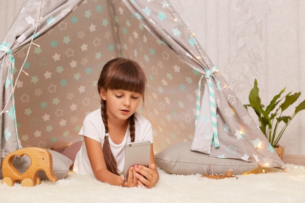 Imagen de una linda niñita de cabello oscuro con coletas usando una camiseta blanca posando en un wigwam en casa viendo dibujos animados mirando un teléfono inteligente con una expresión impresionada concentrada