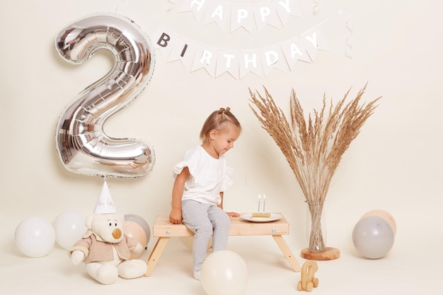 Imagen de una linda niñita rubia con moños de pelo divertidos sentada en un banco de madera cerca del gran número dos hecho de un globo inflable soplando velas en un pastel