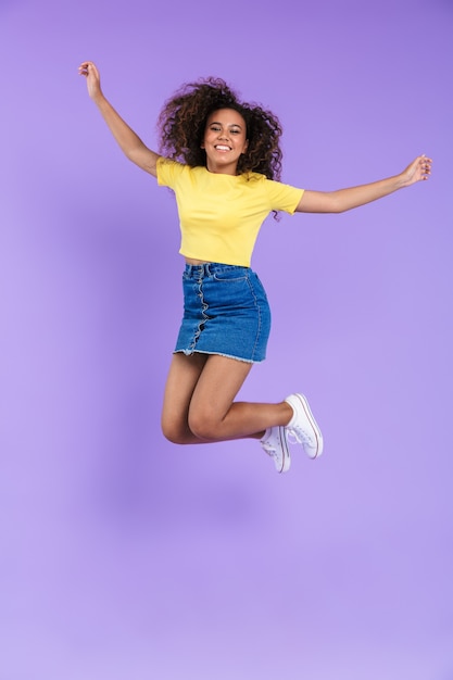 Imagen de longitud completa de la alegre mujer afroamericana con pelo rizado sonriendo y saltando aislado sobre la pared violeta