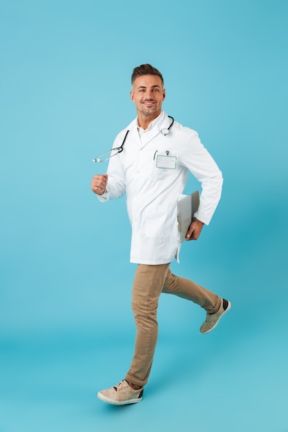 Foto imagen de longitud completa del hombre caucásico vestido con bata médica blanca y estetoscopio caminando, mientras está aislado sobre una pared azul
