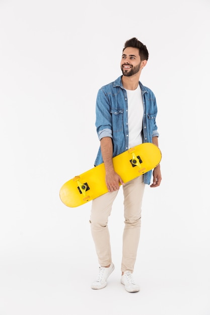 Imagen de longitud completa de hombre feliz sin afeitar sonriendo y de pie con patineta aislado sobre pared blanca