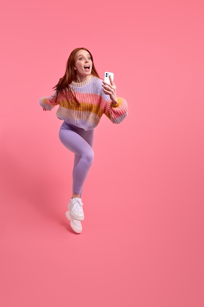 Imagen de longitud completa de mujer gritando sorprendida en ropa casual saltando y apresurarse a tomar una foto en el teléfono inteligente sobre fondo rosa concepto de estilo de vida de personas de emociones humanas