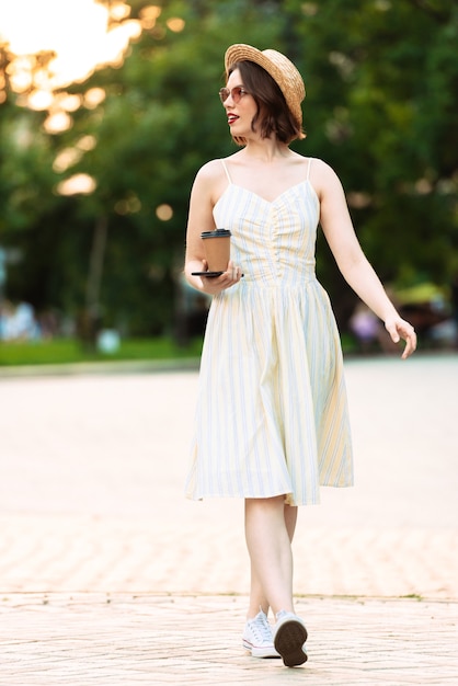 Imagen de longitud completa de mujer sonriente en vestido, sombrero de paja y gafas de sol caminando con una taza de café al aire libre