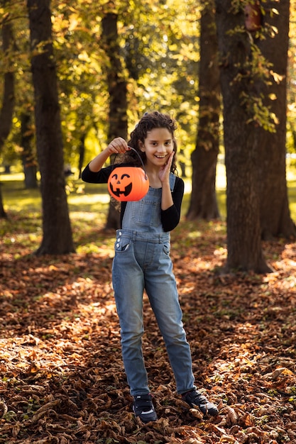 Imagen de longitud completa de una niña bonita con cabello rizado, sosteniendo un caldero naranja de halloween en su mano, sobre un fondo de día soleado de otoño.
