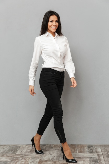 Imagen de longitud completa de sonriente mujer de negocios con camisa blanca y pantalón negro caminando y mirando a un lado, aislado sobre fondo gris