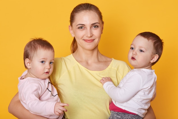 Imagen de madre con hijos, dos hijas en ropa casual, hermosa mujer joven con pequeños gemelos de pie en estudio fotográfico aislado sobre amarillo. Las chicas están interesadas en posar con mami.