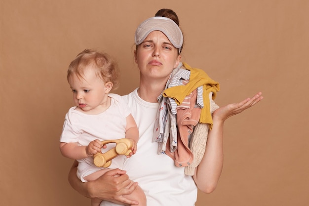 Imagen de una madre joven con un niño pequeño en los brazos extendiendo el brazo a un lado con una expresión facial cansada y molesta que la mujer está exhausta tiene mucho trabajo posando aislada sobre un fondo marrón