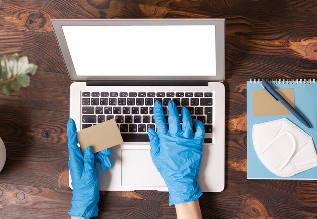 Imagen de manos en guantes médicos sosteniendo una tarjeta de crédito y escribiendo en una computadora portátil. Vista superior.