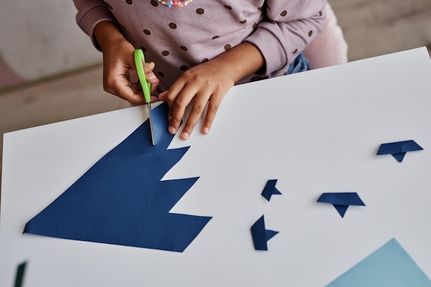 Imagen de manos de una niña afroamericana cortando pequeños barcos de papel azul oscuro mientras está sentada junto a un des