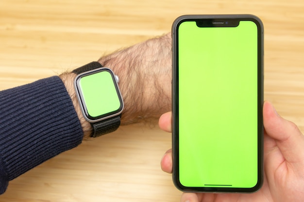 Imagen de manos sosteniendo teléfono móvil negro con pantalla verde y reloj inteligente electrónico en la muñeca.
