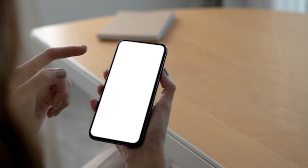 Imagen de maqueta en blanco teléfono celular con pantalla blanca. Mano de mujer sosteniendo mensajes de texto usando el móvil en el escritorio en la oficina en casa.