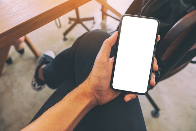 Imagen de la maqueta de la mano de un hombre sosteniendo un teléfono móvil negro con pantalla en blanco con una taza de café en la mesa del café