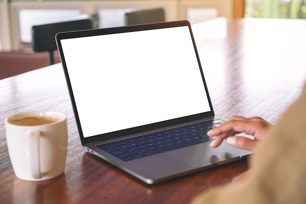Imagen de maqueta de la mano de una mujer usando y tocando en el panel táctil de la computadora portátil con pantalla de escritorio blanca en blanco con una taza de café en la mesa de madera