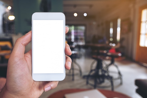 Foto imagen de maqueta de una mano sosteniendo un teléfono móvil blanco con pantalla de escritorio en blanco en la cafetería