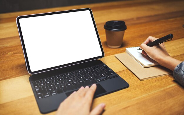 Imagen de maqueta de una mano usando y tocando el panel táctil de la tableta con pantalla de escritorio en blanco en blanco como computadora PC mientras escribe y trabaja, taza de café en la mesa de madera
