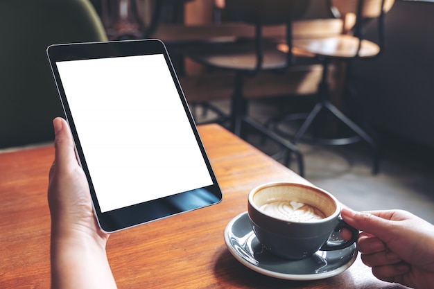 Imagen de maqueta de manos de mujer sosteniendo tablet pc negro con pantalla blanca en blanco con taza de café en la mesa de madera en la cafetería