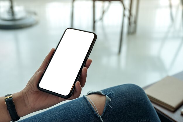 Imagen de maqueta de manos de mujer sosteniendo teléfono móvil negro con pantalla en blanco horizontalmente en café