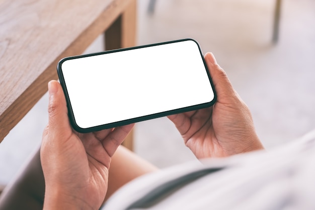 Foto imagen de maqueta de manos de mujer sosteniendo teléfono móvil negro con pantalla en blanco horizontalmente en café