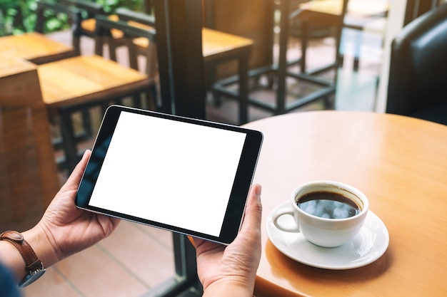 Imagen de maqueta de manos sosteniendo tablet pc negro con pantalla blanca en blanco con taza de café en la mesa de madera