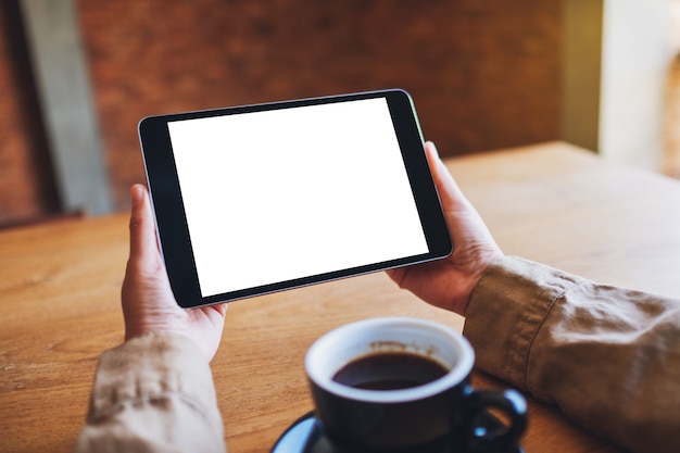 Imagen de maqueta de manos sosteniendo tablet pc negro con pantalla blanca en blanco con taza de café en la mesa de madera