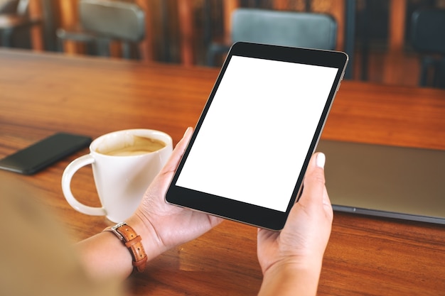 Imagen de maqueta de manos sosteniendo tablet pc negro con pantalla blanca en blanco con taza de café en la mesa de madera
