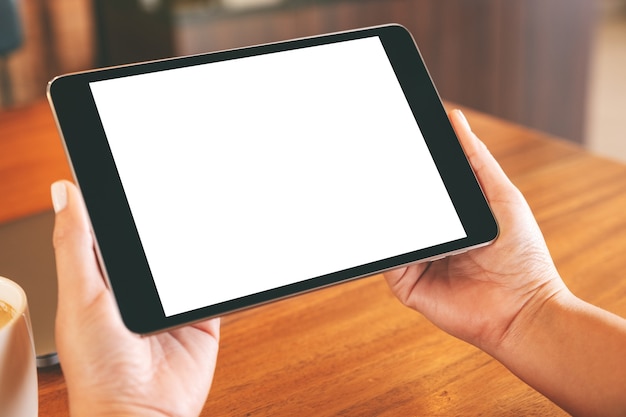Imagen de maqueta de manos sosteniendo tablet pc negro con pantalla en blanco con laptop y taza de café en mesa de madera