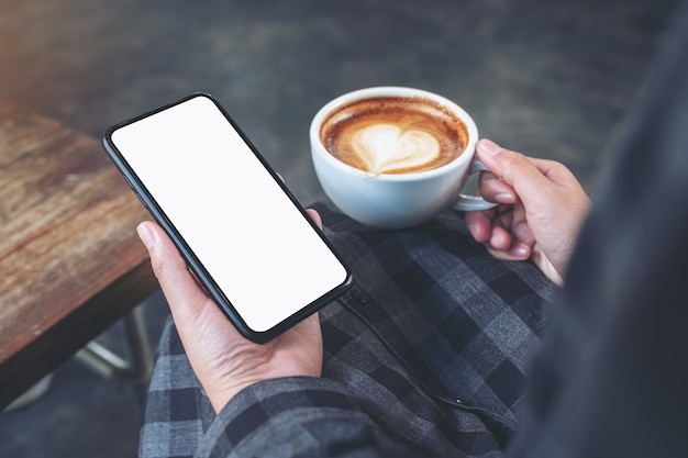 Imagen de maqueta de manos sosteniendo un teléfono móvil negro con pantalla en blanco mientras bebe café en la cafetería