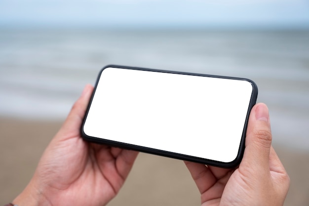 Imagen de maqueta de manos sosteniendo un teléfono móvil negro con pantalla de escritorio en blanco junto al mar