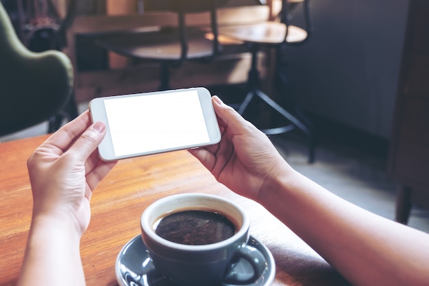 Imagen de maqueta de manos sosteniendo y usando un teléfono móvil blanco con pantalla en blanco horizontalmente para mirar con una taza de café en la mesa de madera