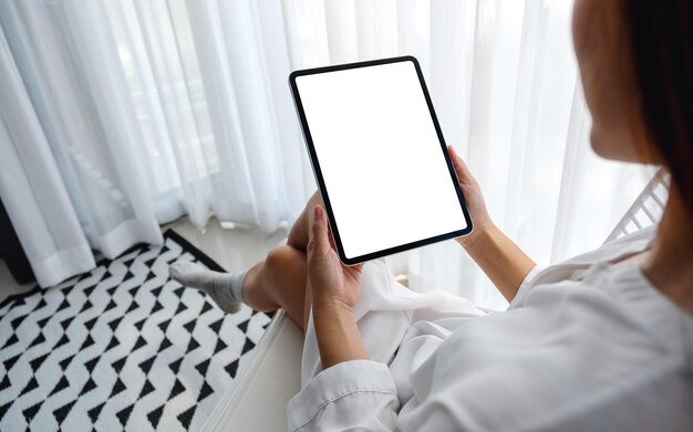 Imagen de maqueta de una mujer que sostiene una tablet pc negra con una pantalla de escritorio blanca en blanco mientras está sentada en el dormitorio con una sensación de relajación por la mañana