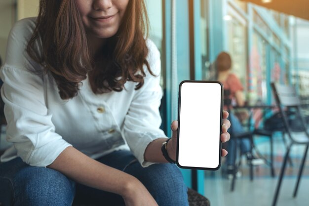 Imagen de maqueta de una mujer sosteniendo y mostrando un teléfono móvil negro con pantalla en blanco en la cafetería