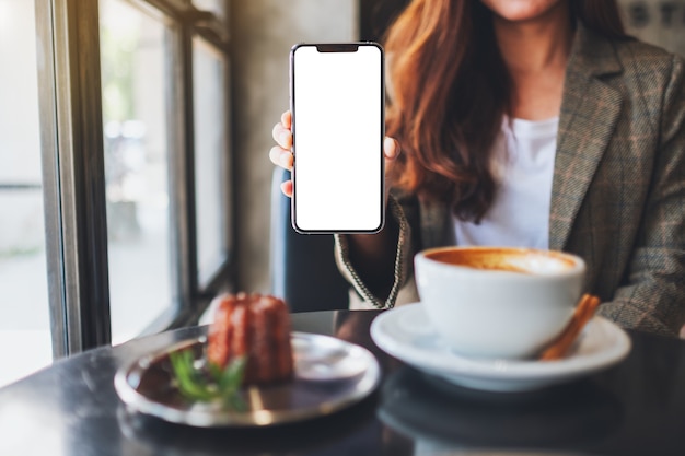 Foto imagen de maqueta de una mujer sosteniendo y mostrando un teléfono móvil negro con pantalla en blanco con una taza de café y un bocadillo en la mesa del café