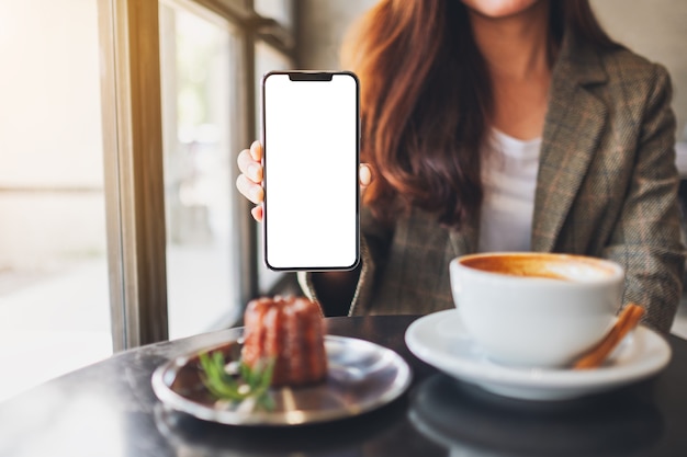 Foto imagen de maqueta de una mujer sosteniendo y mostrando un teléfono móvil negro con pantalla en blanco con taza de café y bocadillos en la cafetería