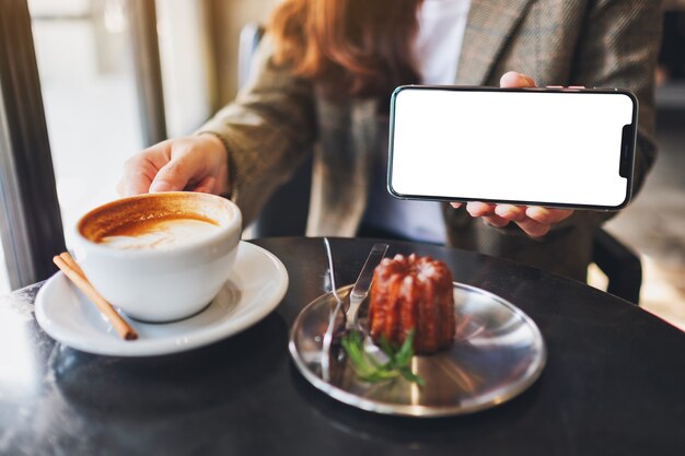 Foto imagen de maqueta de una mujer sosteniendo y mostrando un teléfono móvil negro con pantalla en blanco con taza de café y bocadillos en la cafetería