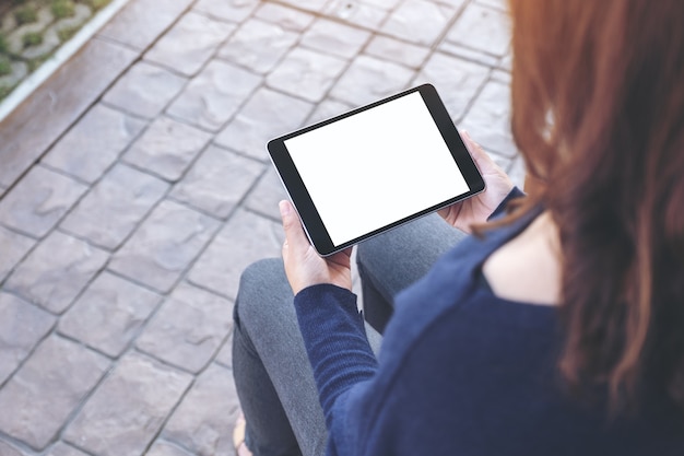 Imagen de maqueta de una mujer sosteniendo una tablet pc negra con pantalla blanca en blanco horizontalmente mientras está sentada al aire libre