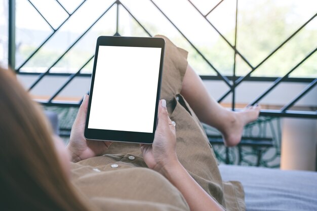 Imagen de maqueta de una mujer sosteniendo una tablet pc negra con una pantalla de escritorio blanca en blanco mientras se acuesta en la sala de estar sintiéndose relajada