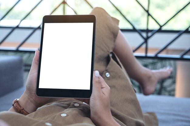 Foto imagen de maqueta de una mujer sosteniendo una tablet pc negra con una pantalla de escritorio blanca en blanco mientras se acuesta en la sala de estar sintiéndose relajada
